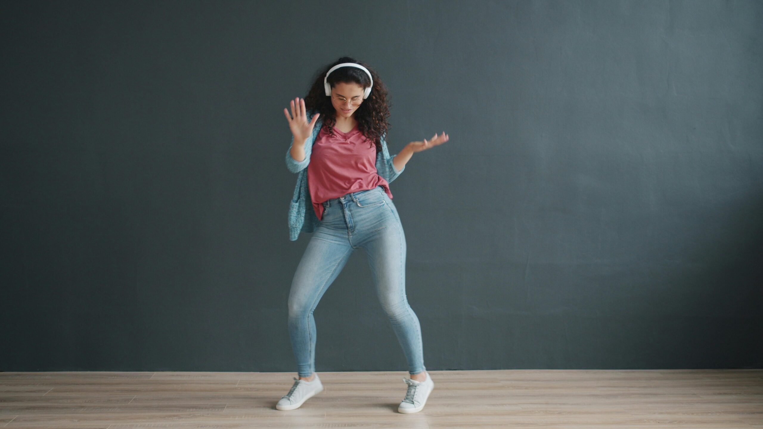 A young woman dances joyfully with headphones indoors against a dark background.