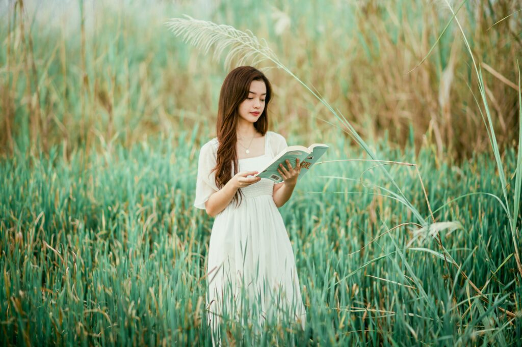 Young elegant ethnic lady in dress reading book among green grass on meadow in summer